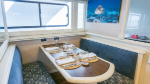 Booth-style dining room table on Galapagos Sky dive boat with blue-padded benches & wooden tabletop set for 4 beside a view window.