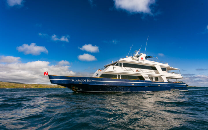 Water-level view of Galapagos Sky dive boat with blue hull & 2 white upper decks, sitting in calm water beside a green island.
