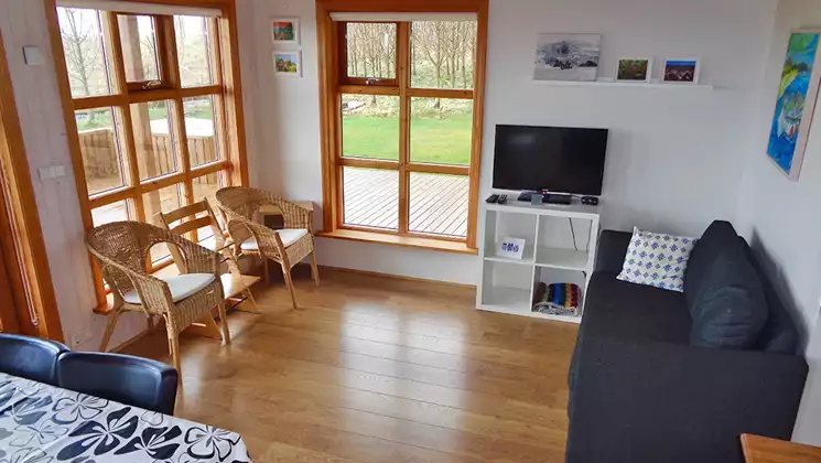 Small living room of a cabin at Hali Country Hotel in Iceland with large windows, blue couch, TV, 2 chairs & wood floor.