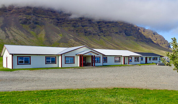 Long single-story white building with metal roof sits by grass & gravel lot backed by verdant cliffs at Hali Country Hotel.