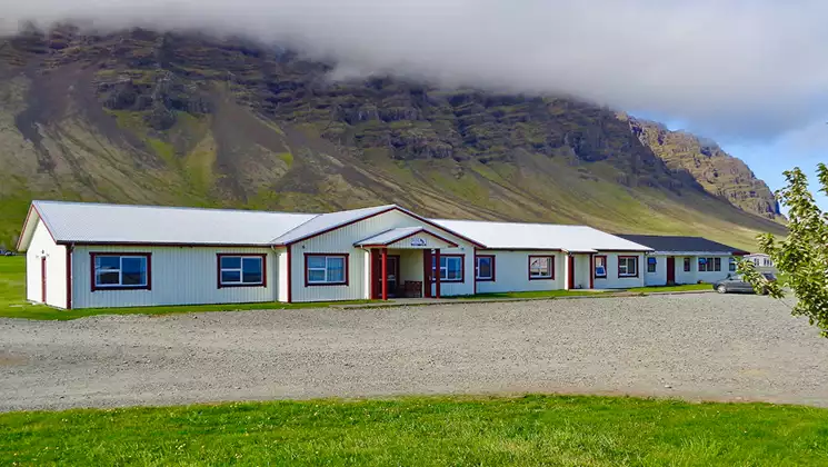 Long single-story white building with metal roof sits by grass & gravel lot backed by verdant cliffs at Hali Country Hotel.