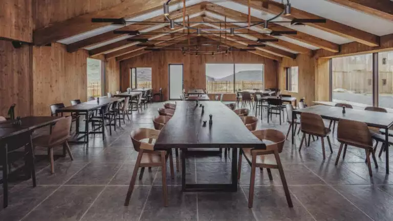 Dining room at Iceland's Highland Base Hotel with long wood banquet tables & modern chairs, high ceiling & walls of windows.