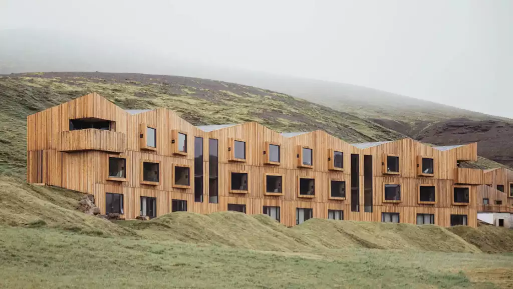 Long, modern, 3-story wooden building with protruding windows backed by green hills at Highland Base Hotel in Iceland.
