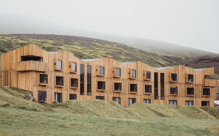 Long, modern, 3-story wooden building with protruding windows backed by green hills at Highland Base Hotel in Iceland.