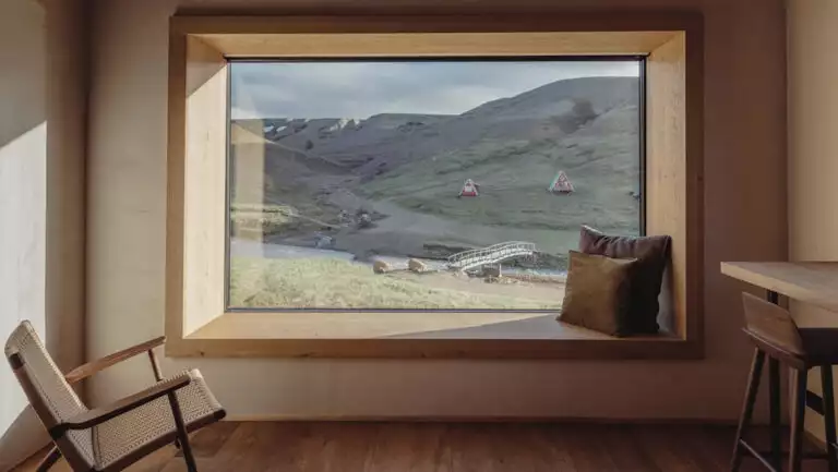 Guest room seating area at Highland Base Hotel in Iceland with woven chair, large bay window, wood bar & tall leather barstool.