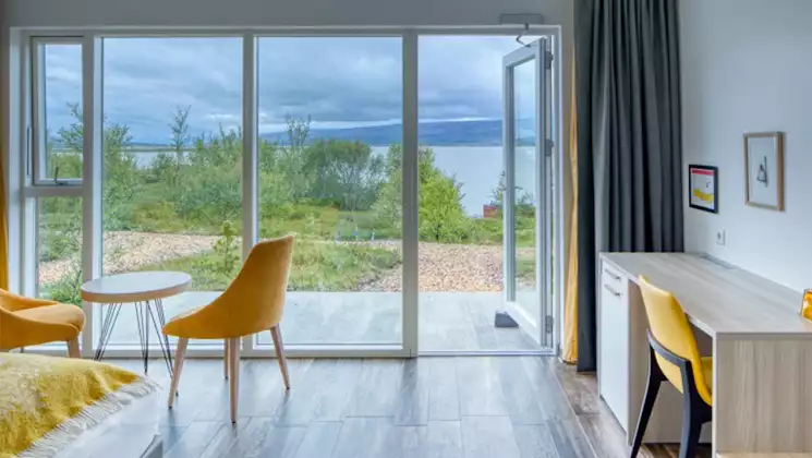 Bright guest room at Iceland's Hotel 1001 Nott with light wood floors & desk, yellow chairs by wall of windows & private patio.