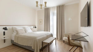 Guest room at Hotel Budir in Iceland with double bed in white linens, minimalist desk & chair, flatscreen TV & curtained window.