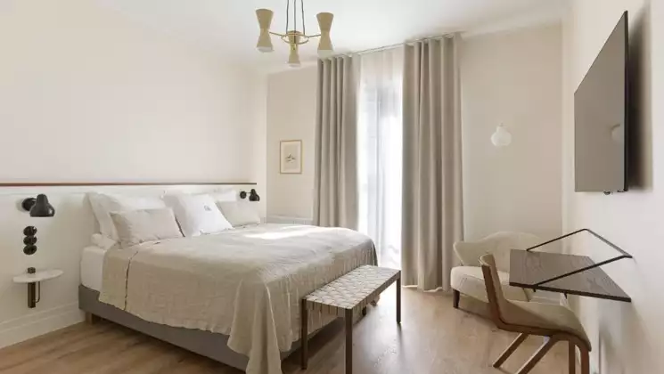 Guest room at Hotel Budir in Iceland with double bed in white linens, minimalist desk & chair, flatscreen TV & curtained window.