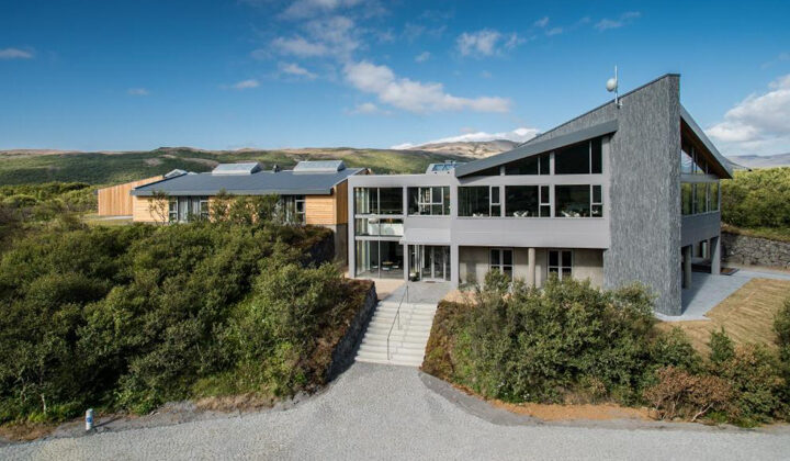 Aerial view of Hotel Husafell in Iceland with modern design of wood, cement, steel & large windows amid green hills & blue sky.