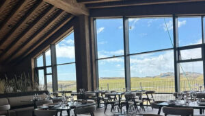 High-ceiling dining room at Hotel Jokulsarlon in Iceland with modern wood tables & wall of windows looking onto green hills.