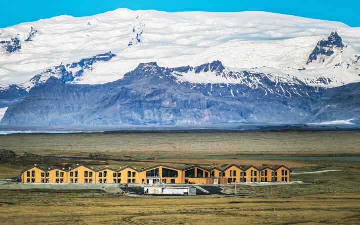 Long building with wood exterior & large glass windows in grass under snowy mountains at Hotel Jokulsarlon in Iceland.