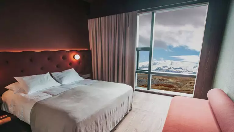 Guest room at Hotel Jokulsarlon in Iceland with double bed in white linens & red velvet headboard, pink couch & view wall.