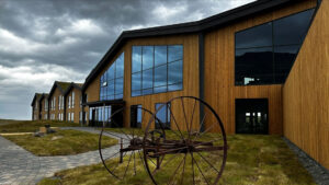 Wood exterior with large glass windows & rusty antique farm equipment in grass under clouds at Hotel Jokulsarlon in Iceland.