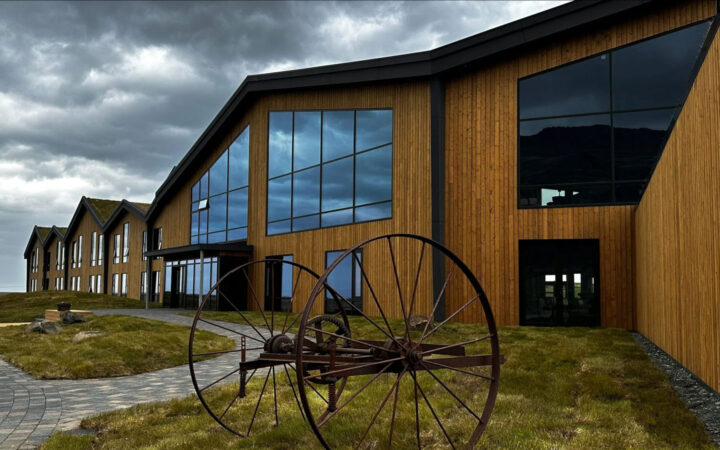 Wood exterior with large glass windows & rusty antique farm equipment in grass under clouds at Hotel Jokulsarlon in Iceland.