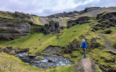 Woman in blue coat stands on overlook with bright green volcanic hills surrounding & river flowing below, in west Iceland.