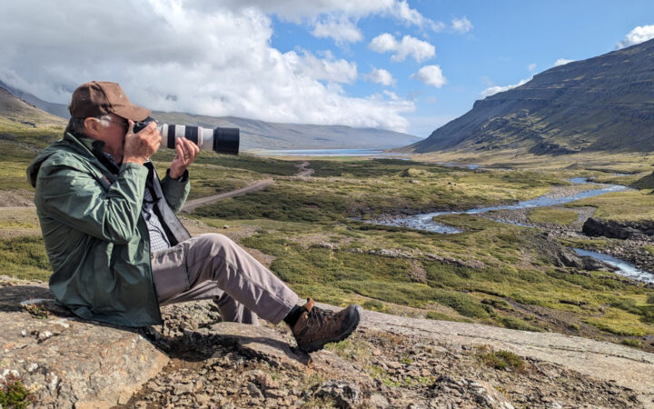 Man in green jacket holds large camera & photographs surrounding green hills & flowing rivers on a sunny day in west Iceland.
