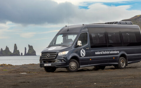 Gray Mercedes Sprinter van sits parked on beach with jagged islands offshore & green volcanic mountains behind, in Iceland.