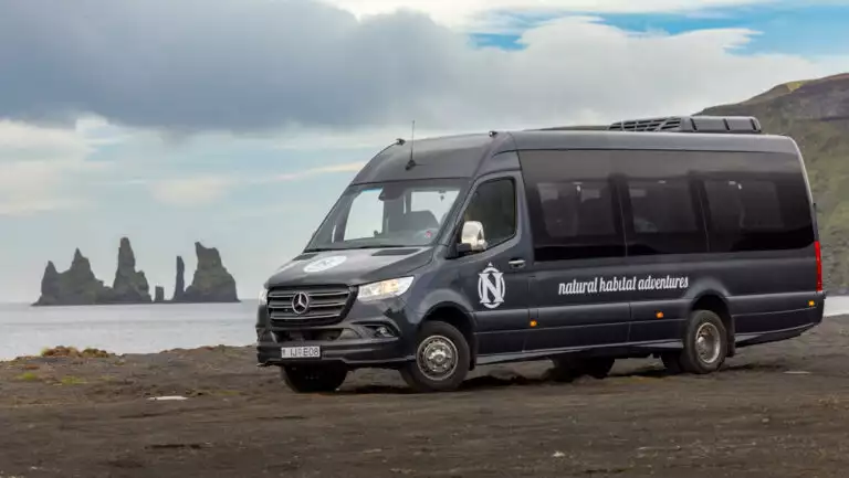 Gray Mercedes Sprinter van sits parked on beach with jagged islands offshore & green volcanic mountains behind, in Iceland.