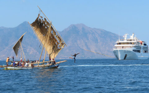 Small white ship sits nearby tiny fishing boat with weathered sails as man jumps off bow throwing a spear in Indonesia.