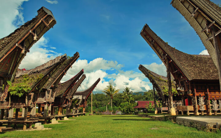 Row of thatched luxury bungalows with tall, pointed roofs facing each other across the grass on a sunny day in Indonesia.