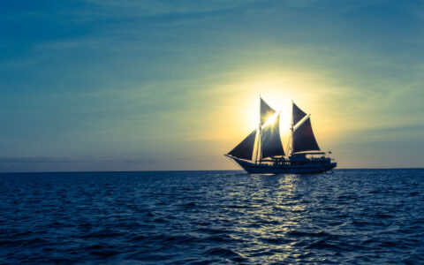 Indonesian sailing ship with sails unfurled cruises while silhouetted by a sunbeam on a calm day.
