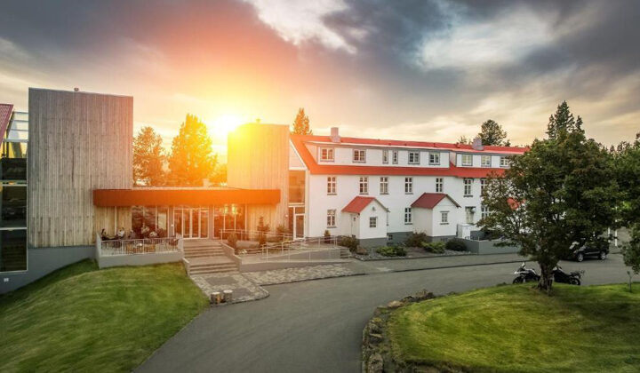 Sun rises over Lake Hotel Egilsstadir with white exterior & red farmhouse roof beside modern stone tower, lawn & trees in Iceland.