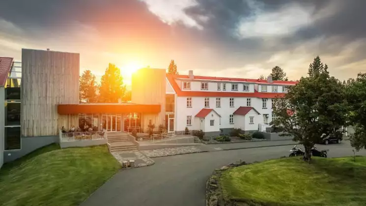 Sun rises over Lake Hotel Egilsstadir with white exterior & red farmhouse roof beside modern stone tower, lawn & trees in Iceland.
