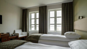 Guest room at Lake Hotel Egilsstadir with 2 twin beds in white linens, wood desk, flatscreen TV, green decor & sunny windows.