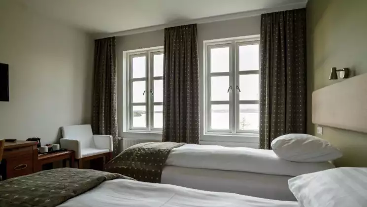 Guest room at Lake Hotel Egilsstadir with 2 twin beds in white linens, wood desk, flatscreen TV, green decor & sunny windows.