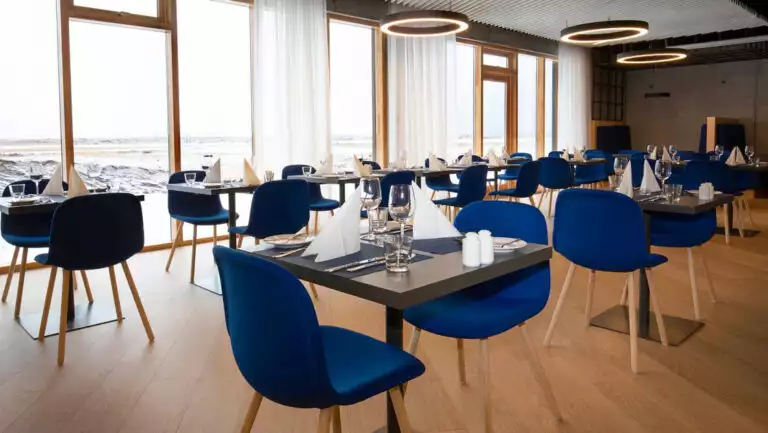 Tindur Restaurant at Landhotel in Iceland, with 2-top tables set for a meal with modern blue chairs, wood floor & wall of windows.