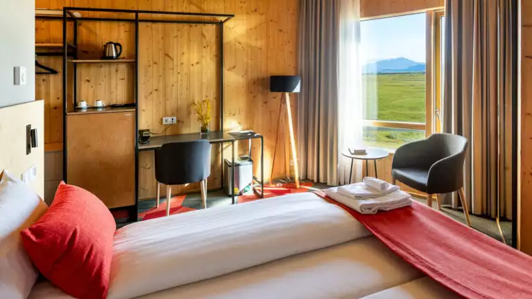 Cozy guest room at Landhotel in Iceland with 2 joined twin beds, wood walls, desk & floor-to-ceiling window to green hills.