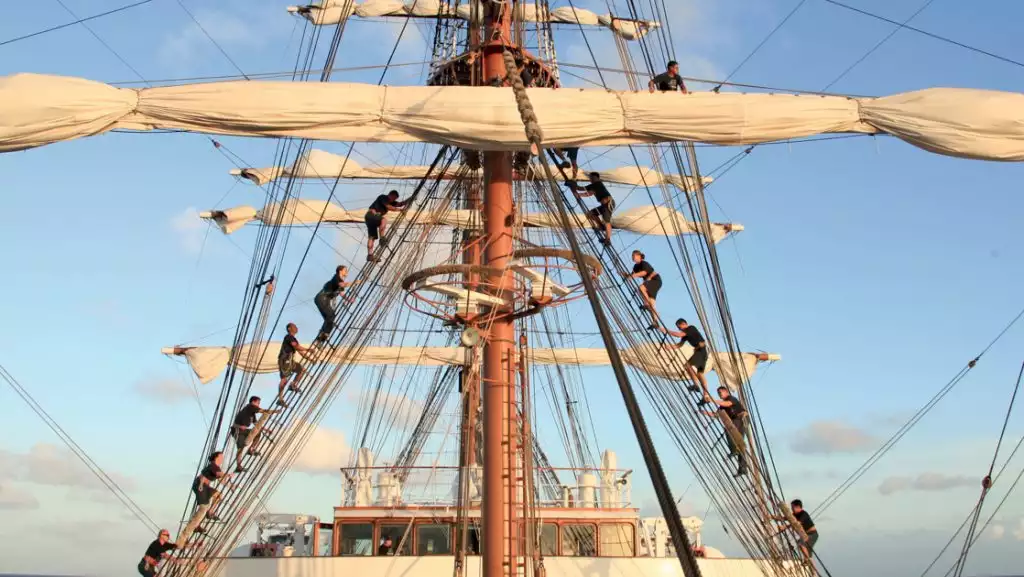 Crew members of Sea Cloud II: Lindblad ship climb the rigging to unfurl the sails with a calm blue sky behind.
