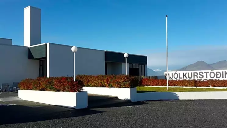 Exterior of Milk Factory Iceland hotel with white industrial walls, red flowers & empty flag pole with mountains in the distance.