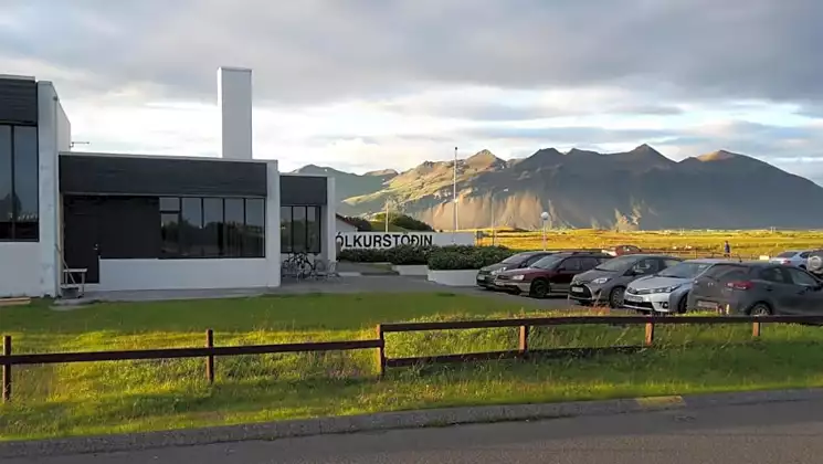 Exterior of Milk Factory Iceland with white industrial walls, tall windows, green grass, cars in parking lot & distant mountains.