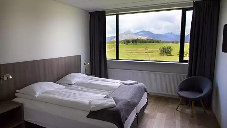 Guest room at Milk Factory hotel with 2 twin beds, laminate wood floor, headboard & bedside tables, modern chair & large window.