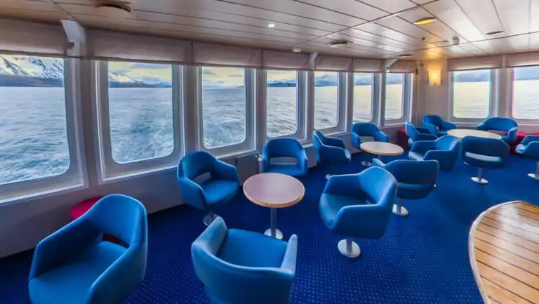 Aft Saloon on Ocean Nova expedition ship with walls of view windows, blue captain's chairs, small tables, blue carpet & wood floor.