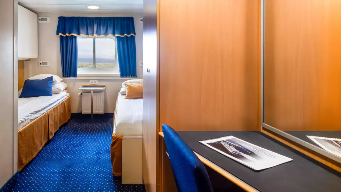 2 lower twin beds in white linens in explorer twin cabin with blue & gold accents, small desk & armoire on Ocean Nova ship.