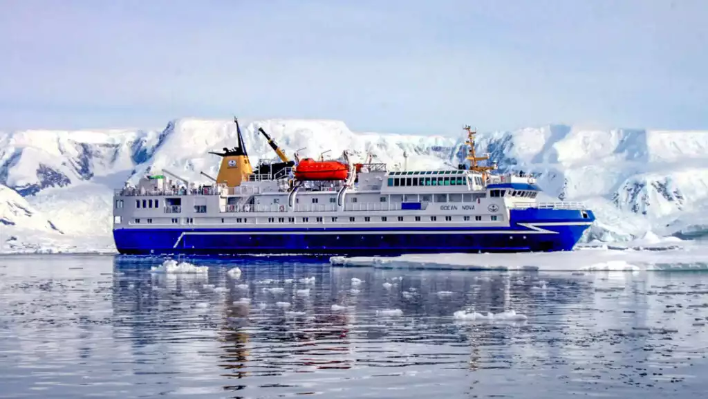 Ocean Nova expedition ship with blue hull, white upper decks & orange lifeboat sits in glassy water beside snowy Antarctic cliffs.