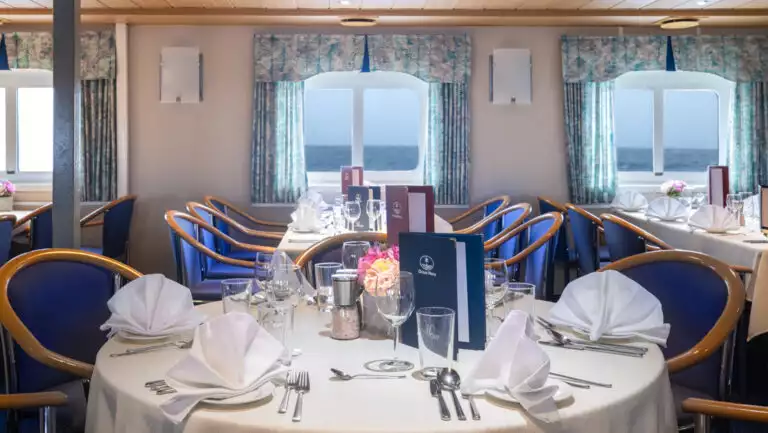 Dining room on Ocean Nova polar ship with view windows & 4- & 6-top tables set in white linens & upscale dinnerware.