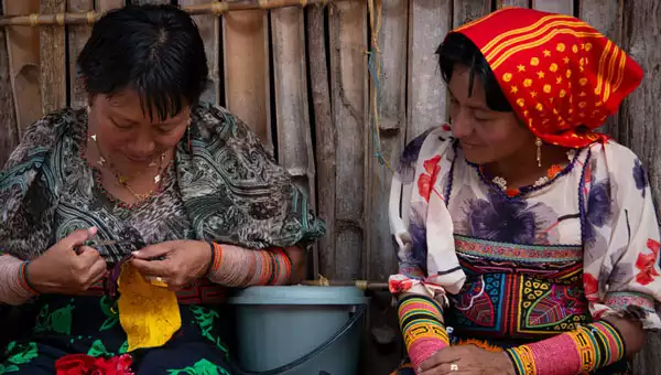 Two women in brightly colored clothes with colored beads covering the forearms sit & sew together, seen on a Panama vacation.