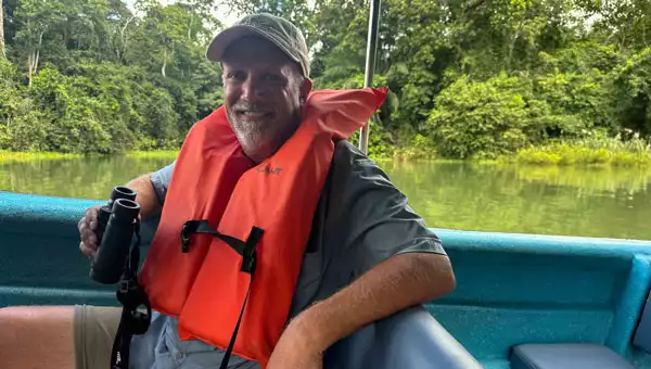 Man in beige hat, short, t-shirt & PFD sits in a boat with binoculars in hand, while drifting among jungle riverbanks on a Panama vacation.