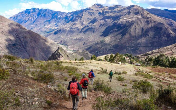 Trekkers on a Peru tour walking down a scenic green mountain toward a village on a sunny day with large mountains in the distance.