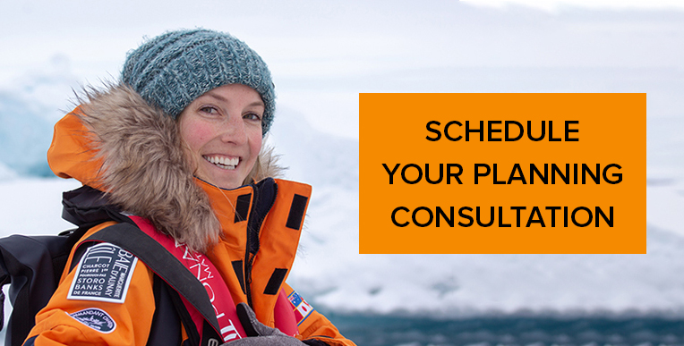 A travel specialist in a beanie and yellow coat with a fur hood seen smiling at the camera in portrait style with a glacier behind her