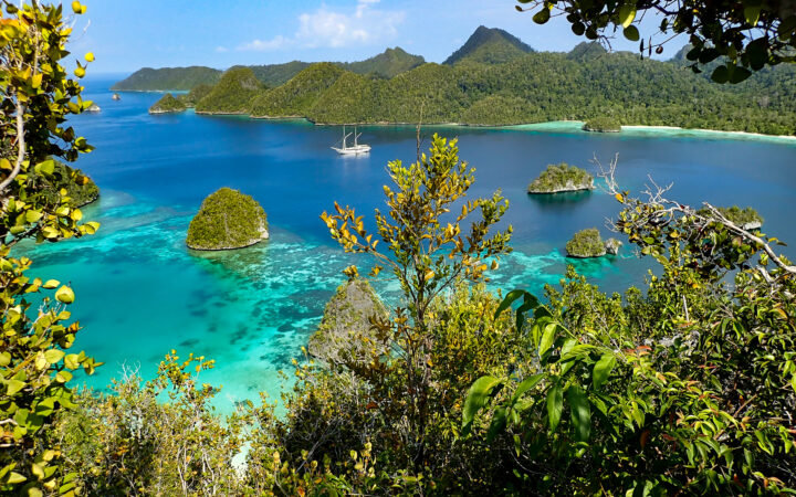 looking down at a beautiful desolate indonesia cove with a small ship anchored in the middle of it and jungle surrounding it.