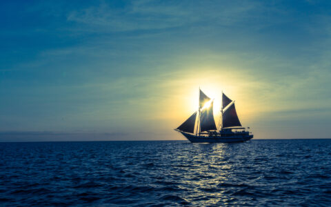 Indonesian sailing ship with sails unfurled cruises while silhouetted by a sunbeam on a calm day.