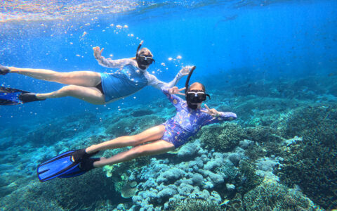 Mother & daughter snorkel in blue sea above colorful corals on a Jewels of Raja Ampat Indonesia sailing cruise.