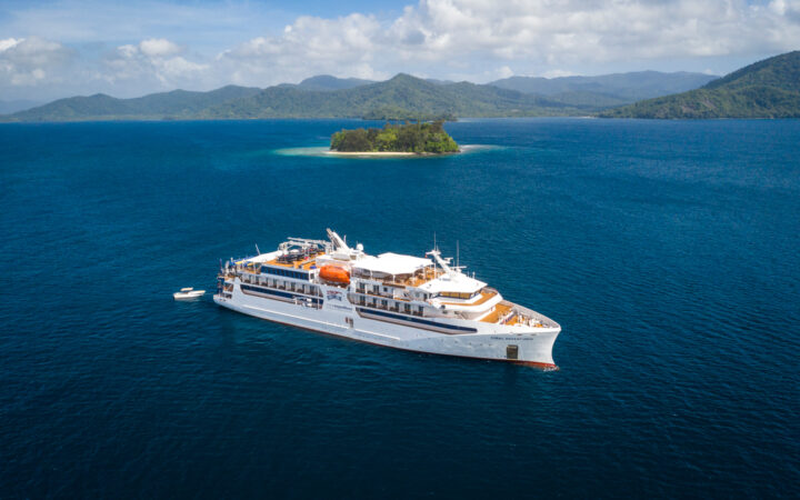 Small white expedition ship with teak decks & white tender vessel behind sits in calm blue sea by tiny white-sand island.