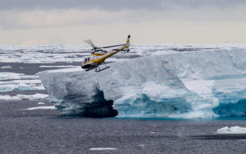 Black, white & yellow helicopter flies above large blue shelf iceberg on the Remote Weddell Explorer Antarctic cruise.