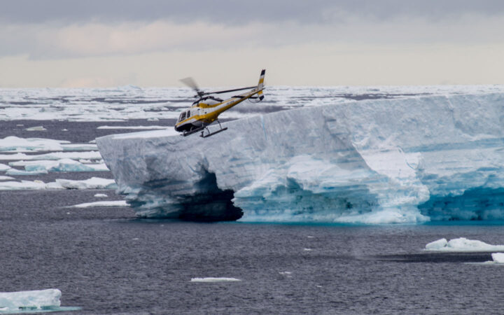 Black, white & yellow helicopter flies above large blue shelf iceberg on the Remote Weddell Explorer Antarctic cruise.