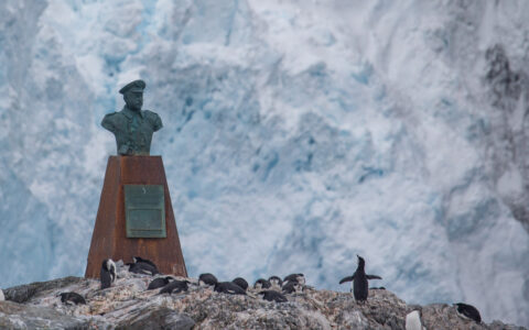Bust of Sir Ernest Shackleton sits atop rusty platform with penguins underneath & large iceberg behind at Point Wild.